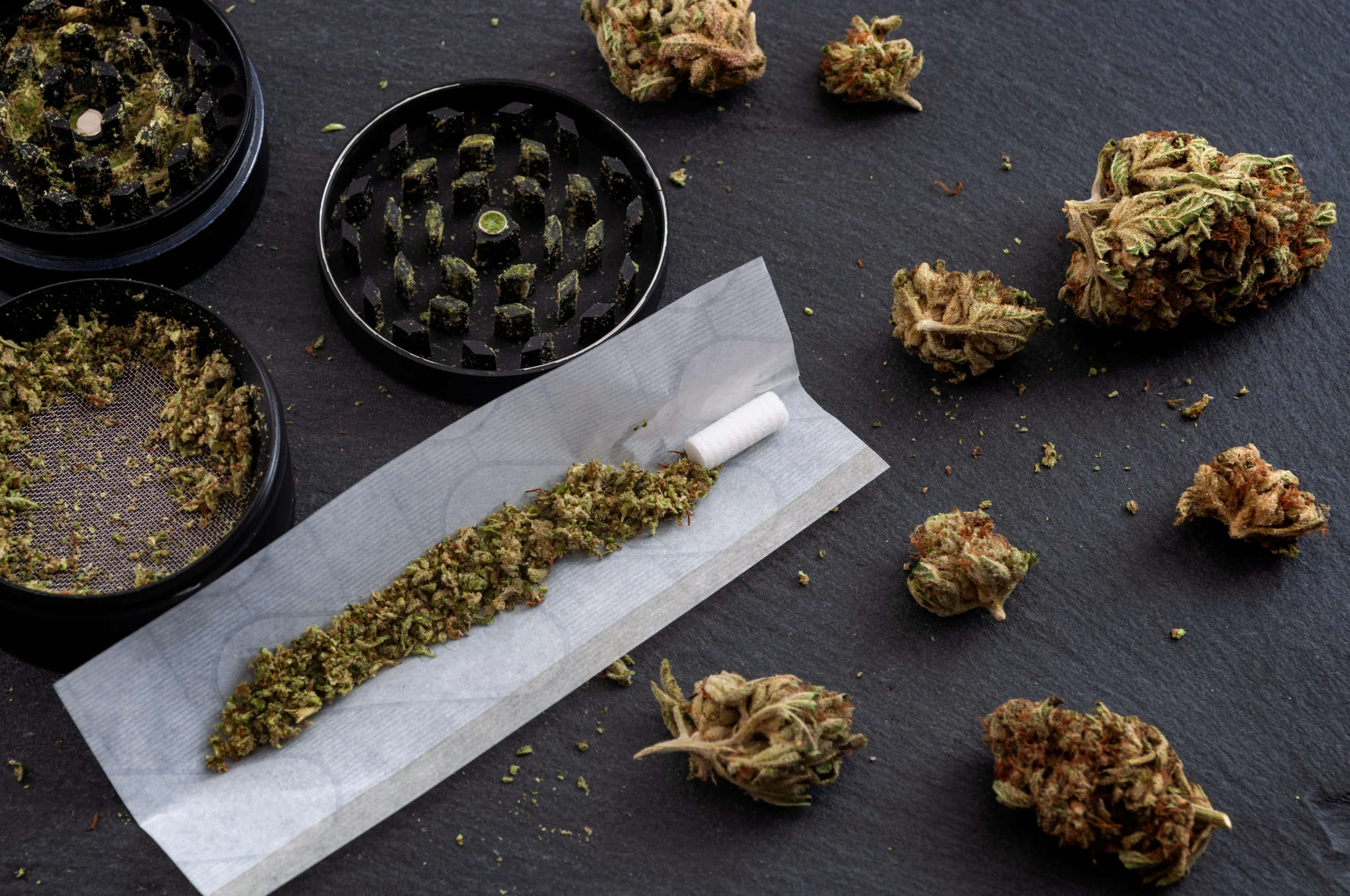 Grinder, loose cannabis flower, and a partially filled rolling paper laid out on a black surface.