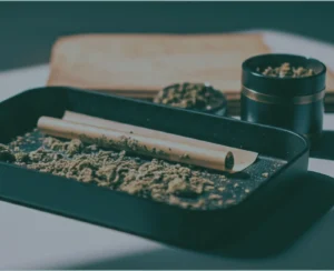 Black rolling tray with partially filled rolling paper, surrounded by ground cannabis and a grinder in the background.