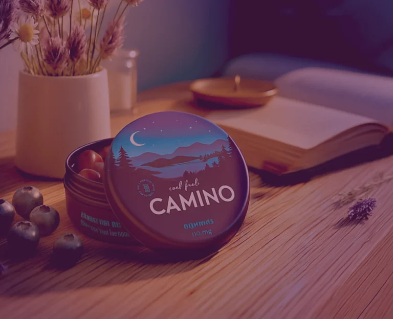 A tin of Camino "Cool Blue" cannabis gummies sits open on a wooden table next to an open book, a candle, and blueberries, evoking a calm evening setting.