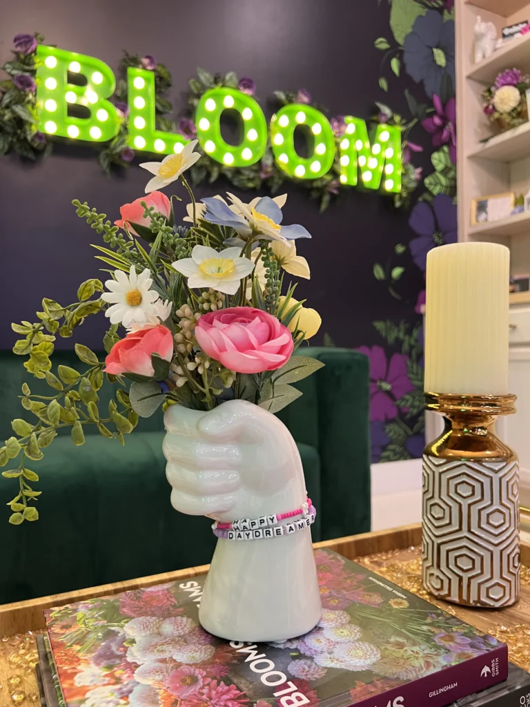 A close-up of a ceramic hand-shaped vase holding a colorful bouquet of artificial flowers, with a beaded bracelet reading “HAPPY DAYDREAMER” around the wrist. The background features a green neon 'BLOOM' sign on a dark floral wall.