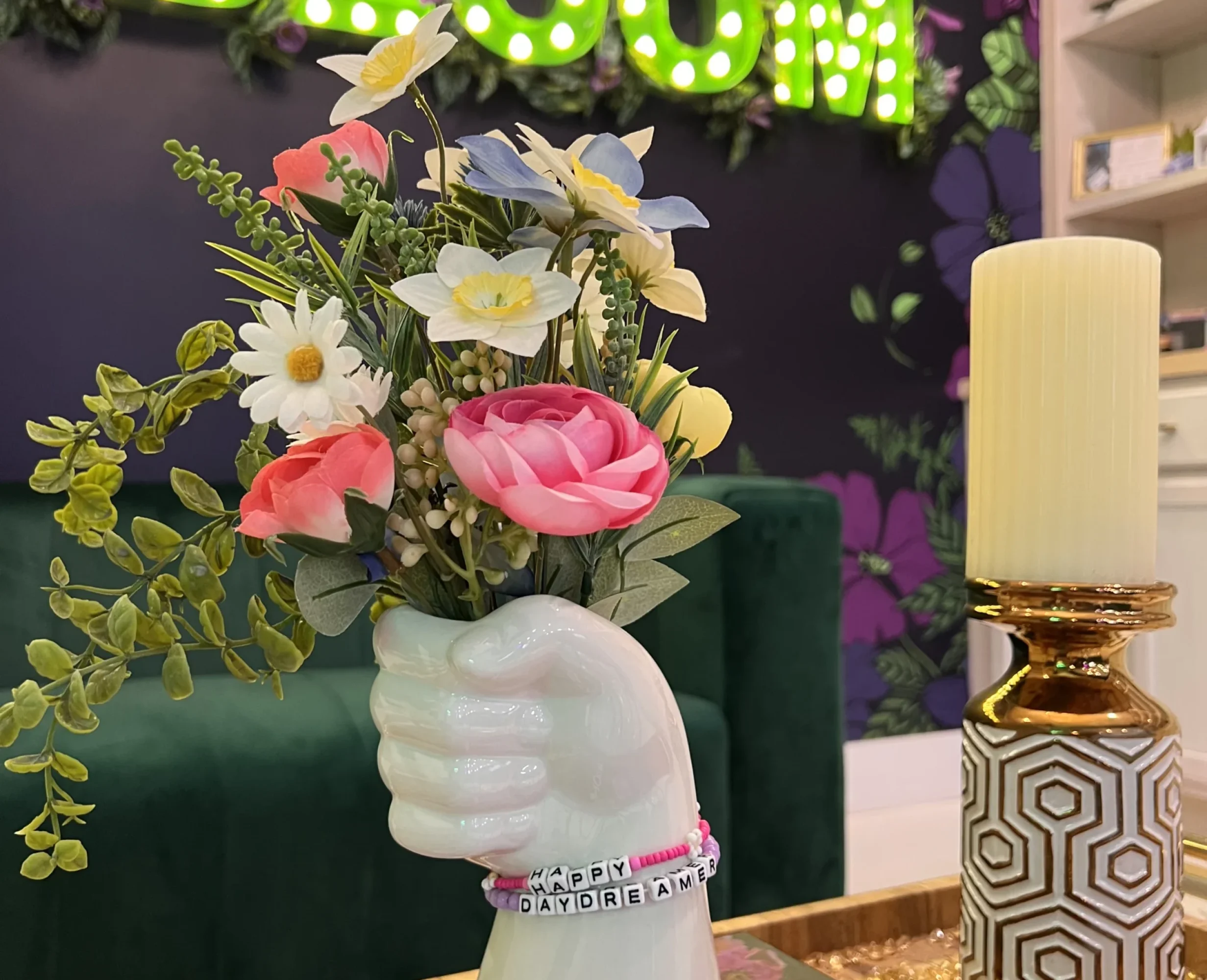A close-up of a ceramic hand-shaped vase holding a colorful bouquet of artificial flowers, with a beaded bracelet reading “HAPPY DAYDREAMER” around the wrist. The background features a green neon 'BLOOM' sign on a dark floral wall.
